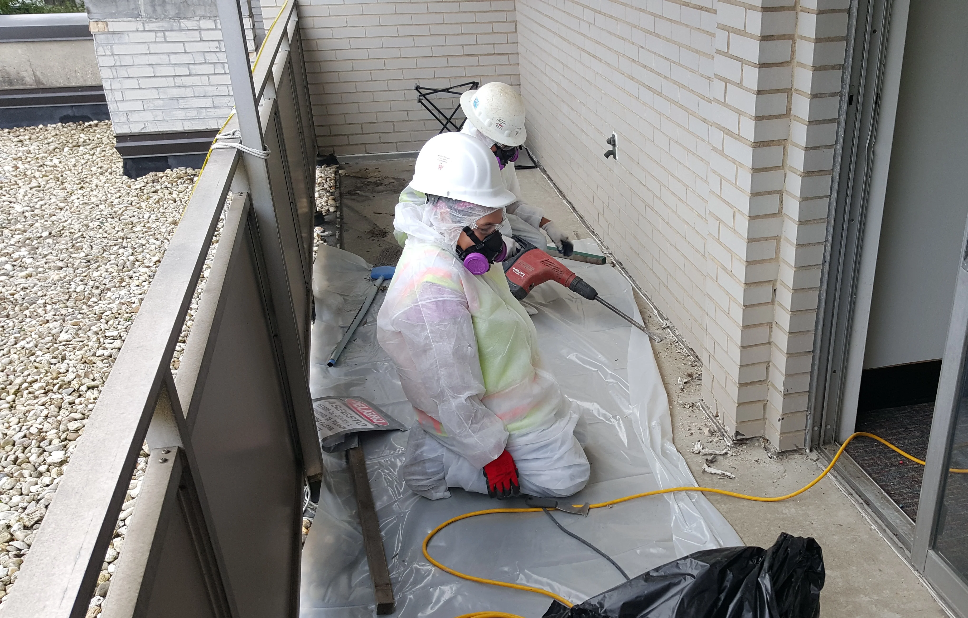 Worker in protective gear removing floor caulk in confined brick utility space